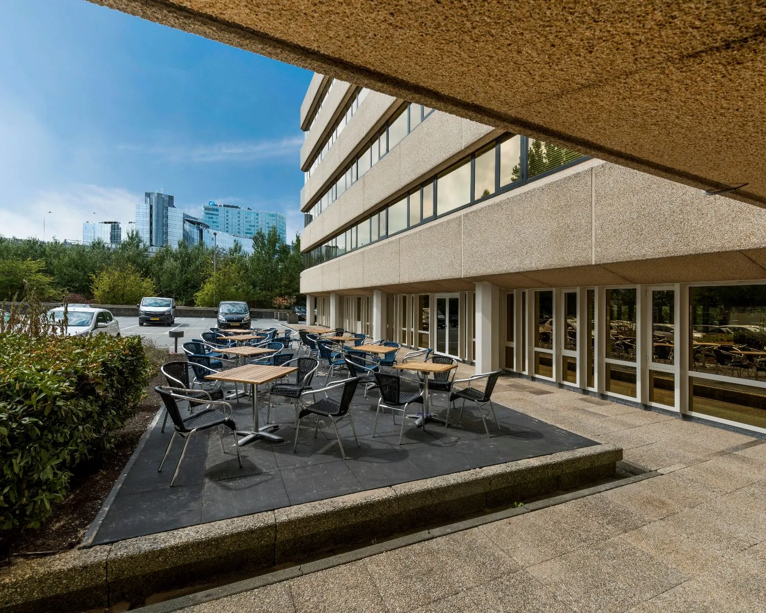 Terras met tafels en stoelen naast een modern kantoorgebouw met uitzicht op hoge gebouwen in de verte.