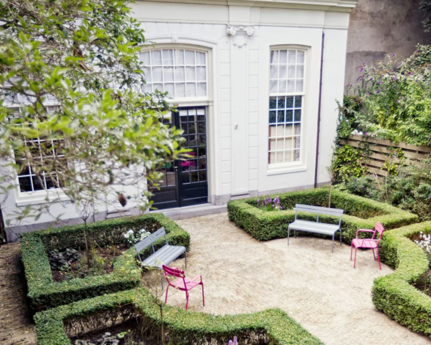 Kleine, symmetrisch aangelegde stadstuin met buxushagen, kleurrijke stoelen en een wit herenhuis op de achtergrond.