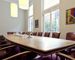 Vergaderruimte met lange houten tafel, leren stoelen, moderne kunst aan de muur en grote ramen met uitzicht op een binnentuin.