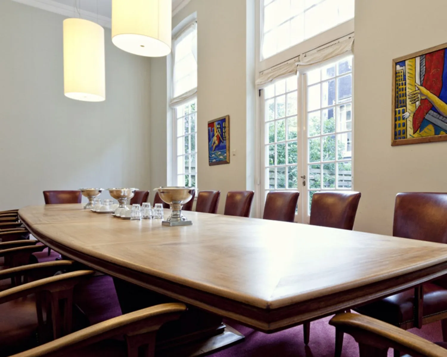 Vergaderruimte met lange houten tafel, leren stoelen, moderne kunst aan de muur en grote ramen met uitzicht op een binnentuin.
