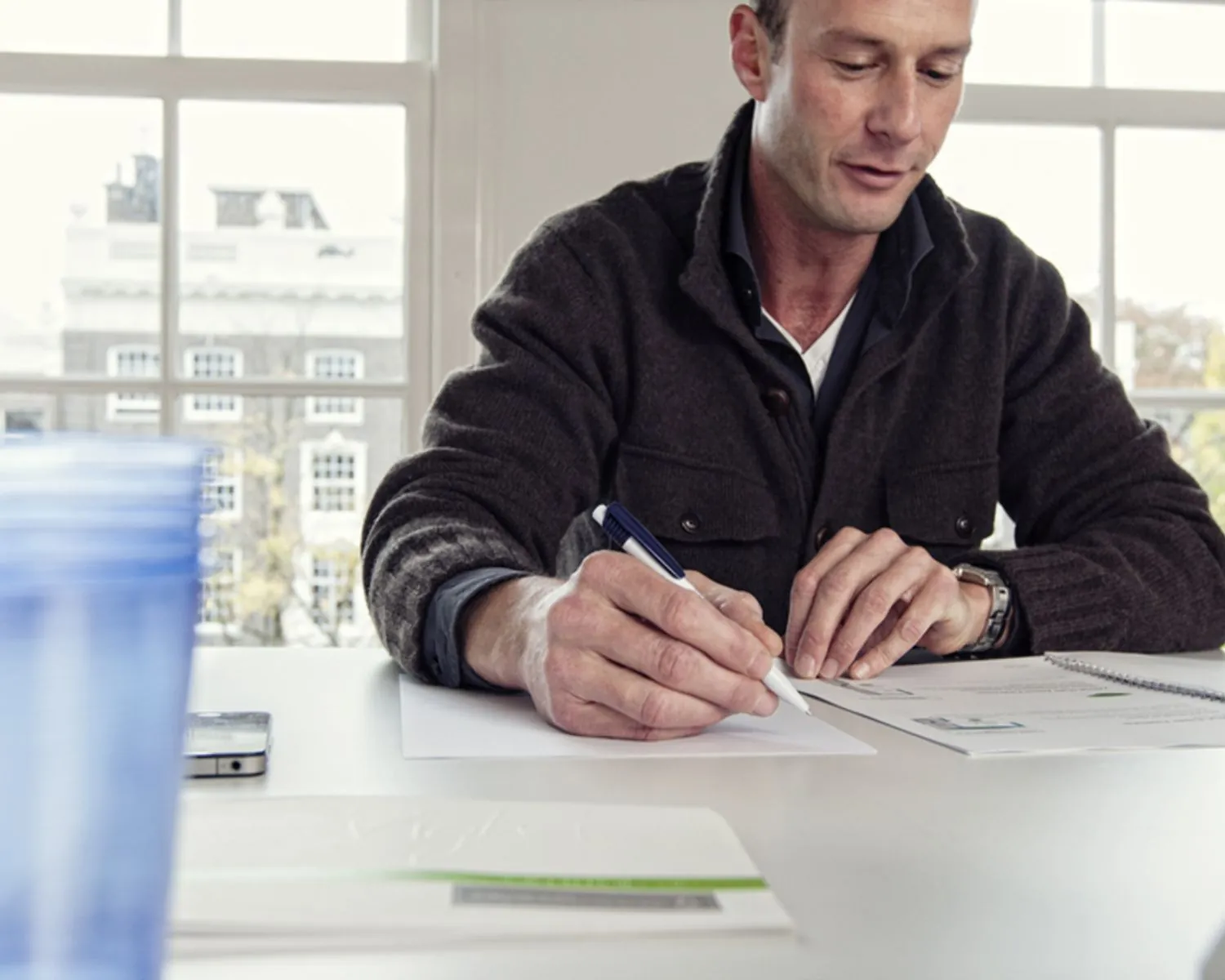 Man schrijft met een pen aan een bureau voor een raam met papieren en een notitieboekje.