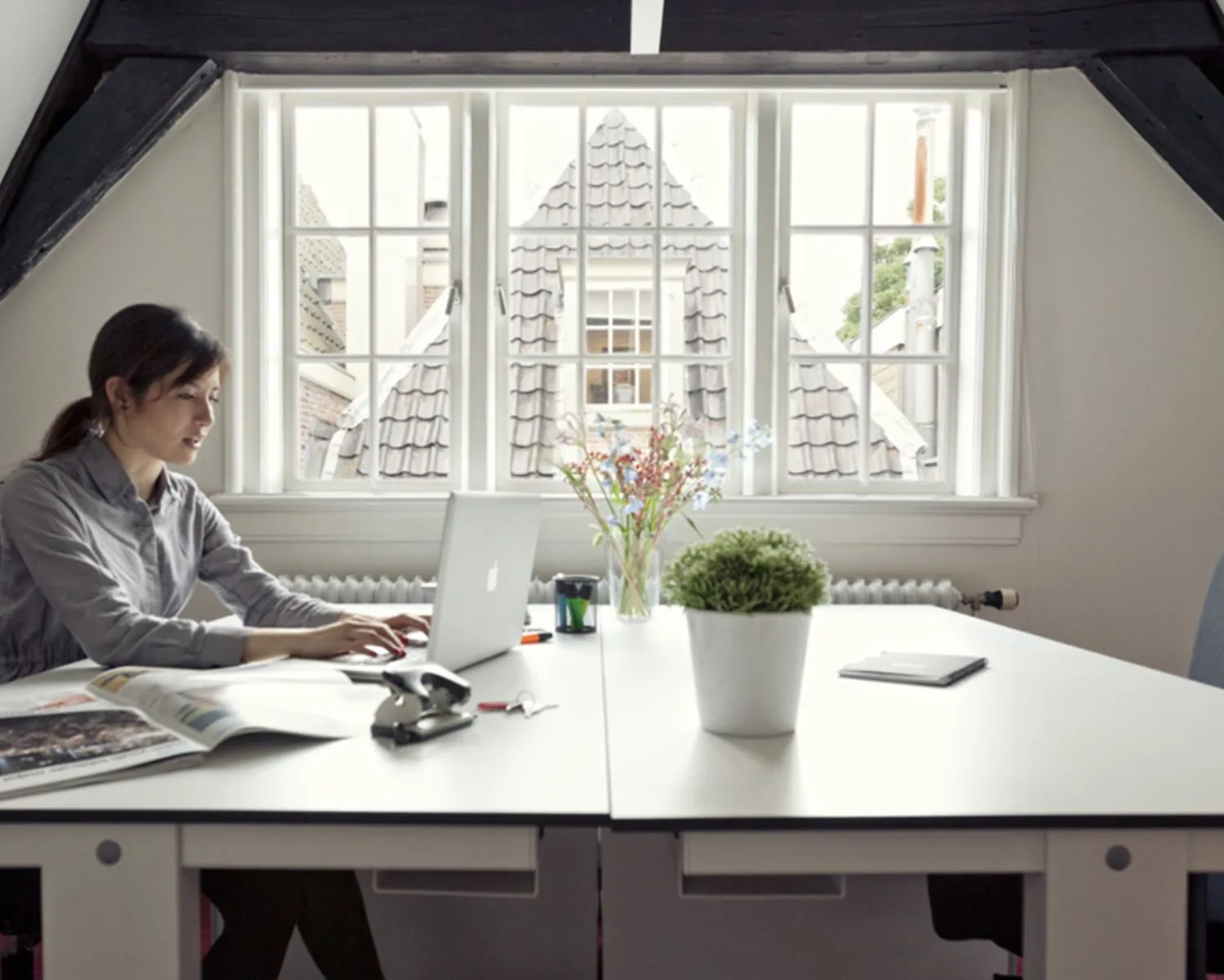 Vrouw werkt op een laptop aan een bureau in een licht kantoor met uitzicht op traditionele dakpannen door het raam.