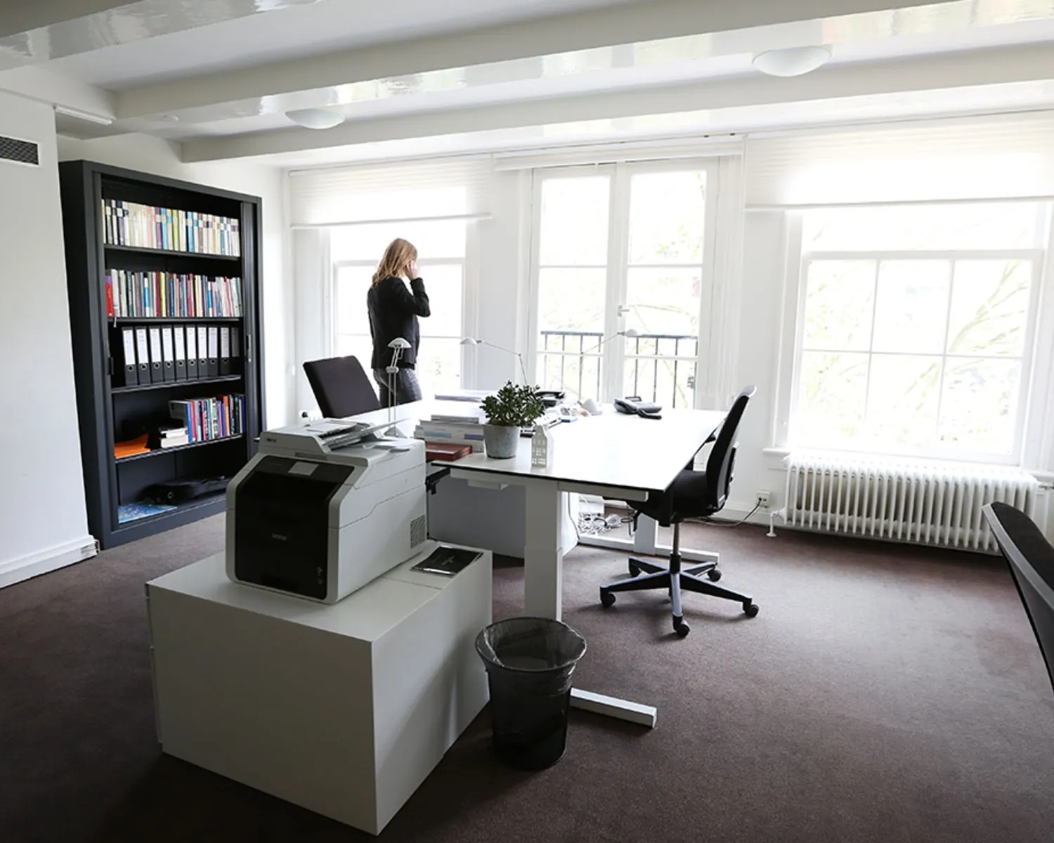 Een moderne, lichte kantoorruimte met bureaumeubilair, een printer en een vrouw die bij het raam telefoneert.