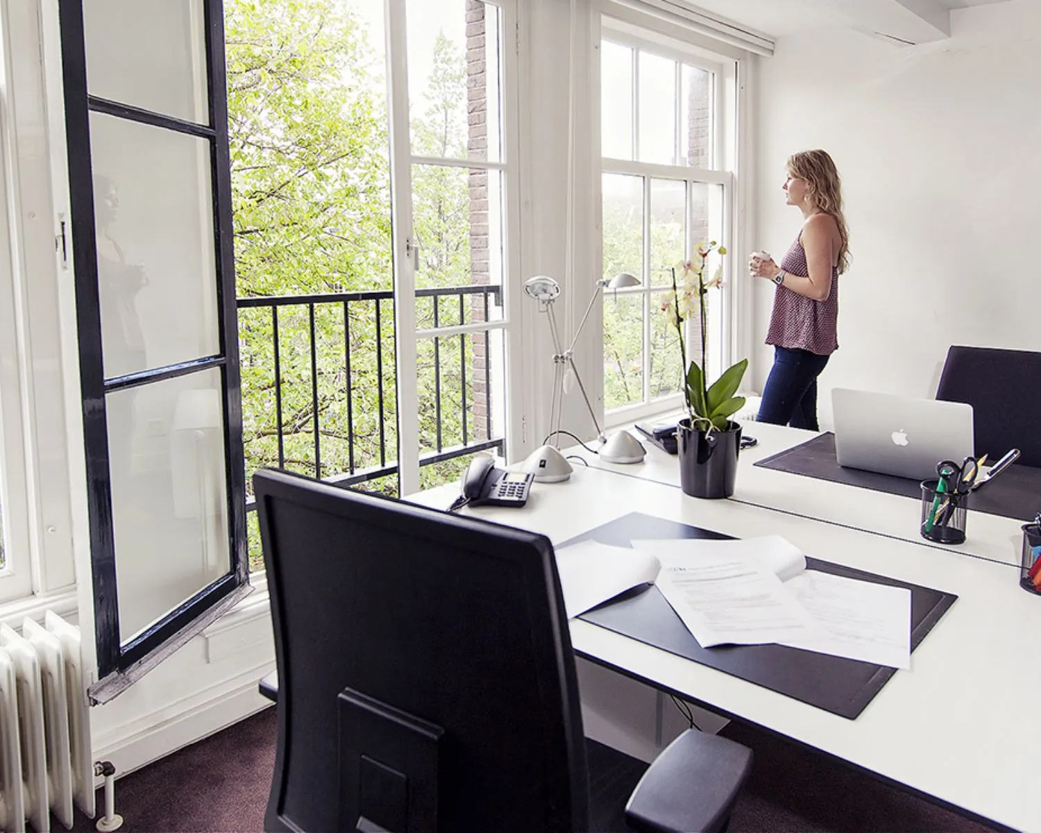 Vrouw met een kopje koffie kijkt uit het raam in een modern, licht kantoor met openstaande ramen en bureaus.