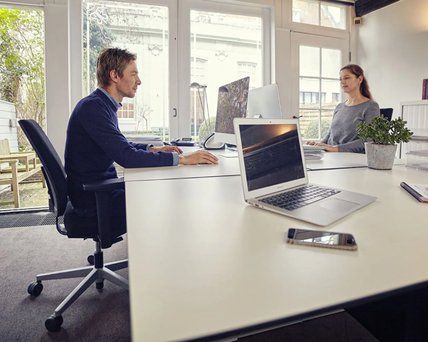 Twee mensen werken tegenover elkaar aan bureaus in een modern en licht kantoor met grote ramen.