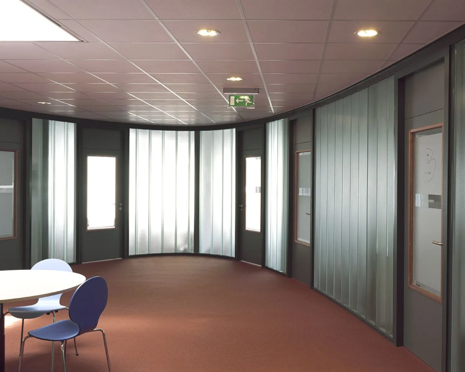 Moderne kantoor- of schoolgang met gebogen glazen wanden, deuren en een tafel met stoelen op roodtapijt.
