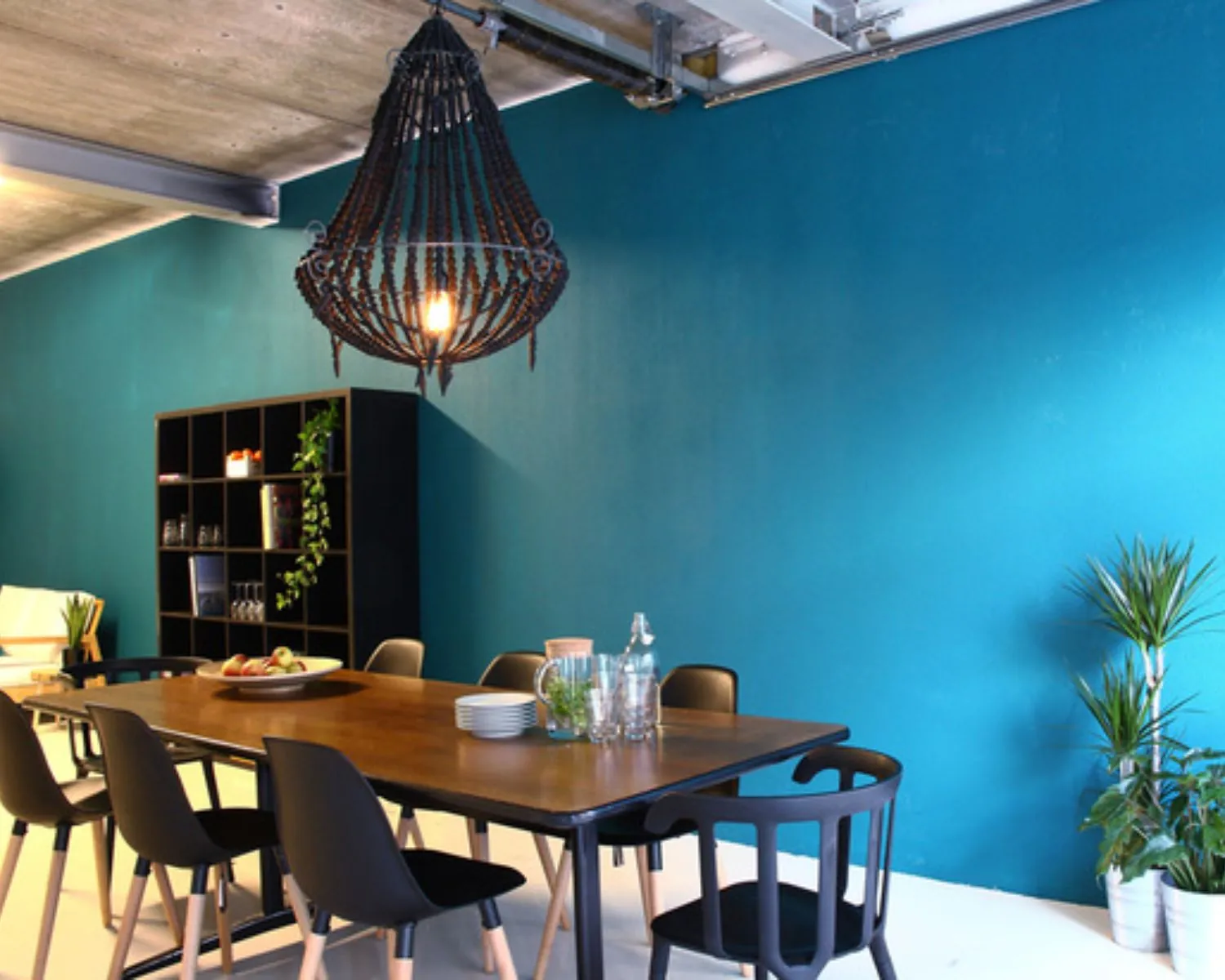 Modern ingerichte eetruimte met houten tafel, zwarte stoelen, opvallende kroonluchter en een turquoise muur.