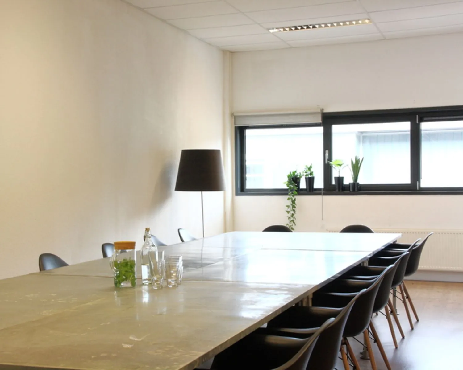 Vergaderruimte met een lange tafel, zwarte stoelen, een staande lamp en planten bij het raam.