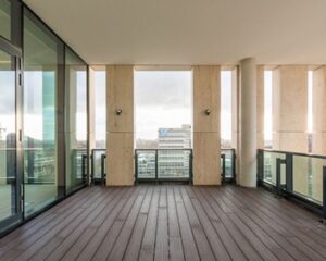 Luxe overdekt balkon met houten vlondervloer, glazen balustrade en uitzicht op moderne stadspanden.