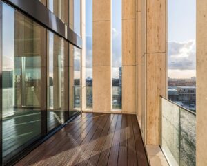 Modern balkon met houten vloer, glazen balustrade en hoge stenen zuilen met uitzicht op de stad.
