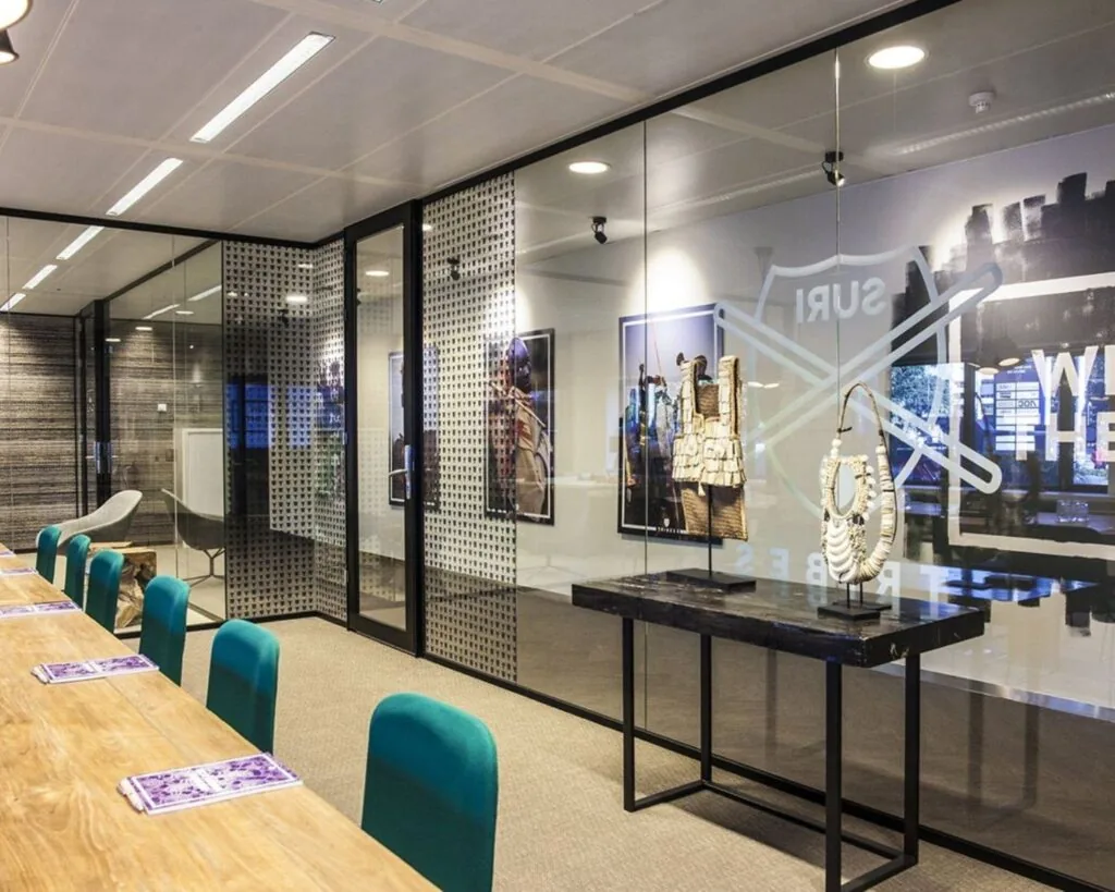 Moderne vergaderruimte met glazen wanden, turquoise stoelen en etnische kunstobjecten aan de muur.