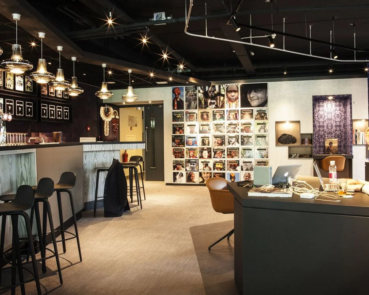 Modern ingerichte kantoorruimte met een bar, hanglampen en een muur vol portretfoto’s van diverse mensen.