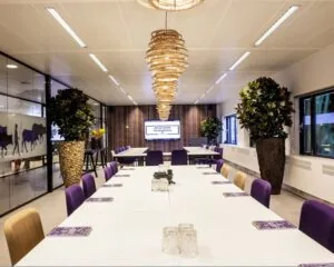 Moderne vergaderruimte met lange witte tafels, paarse stoelen, grote planten en een scherm met de tekst “We go where the business is”.