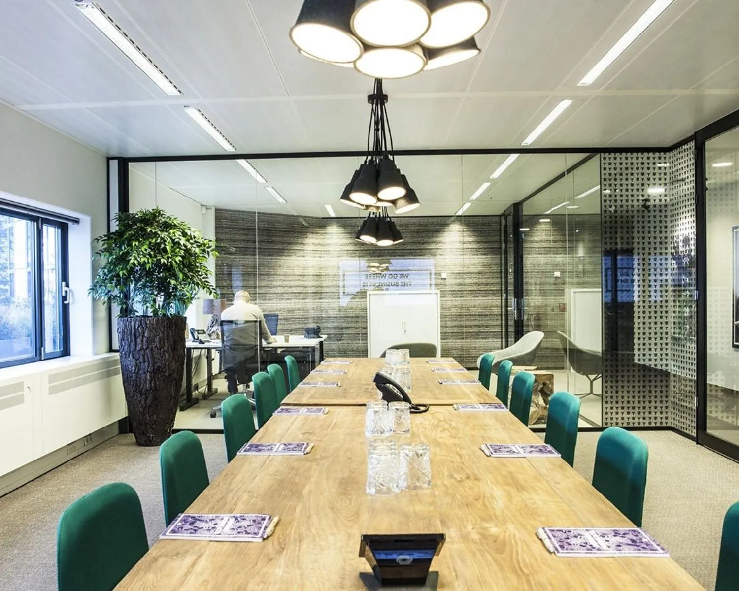 Moderne vergaderruimte met een lange houten tafel, groene stoelen en glazen wanden met uitzicht op een kantoorruimte.