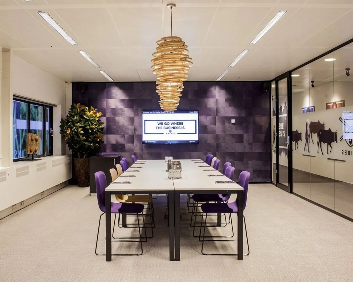Moderne vergaderruimte met een lange tafel, paarse stoelen, een opvallende houten hanglamp en een scherm met de tekst "We go where the business is".