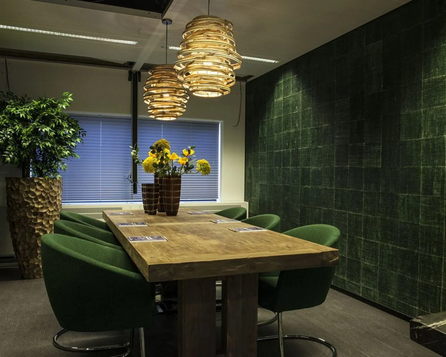Vergaderruimte met een houten tafel, groene stoelen, hanglampen van hout en een groene accentmuur.