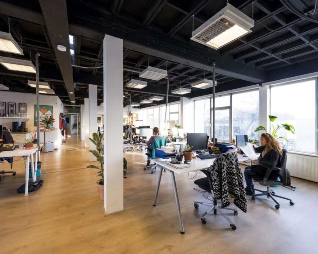 Moderne open kantoorruimte met grote ramen, houten vloer en mensen die aan bureaus werken.