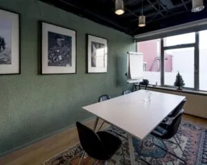 Kleine vergaderruimte met een witte tafel, zes zwarte stoelen, drie zwart-witfoto's aan de muur en een flip-over bij het raam.