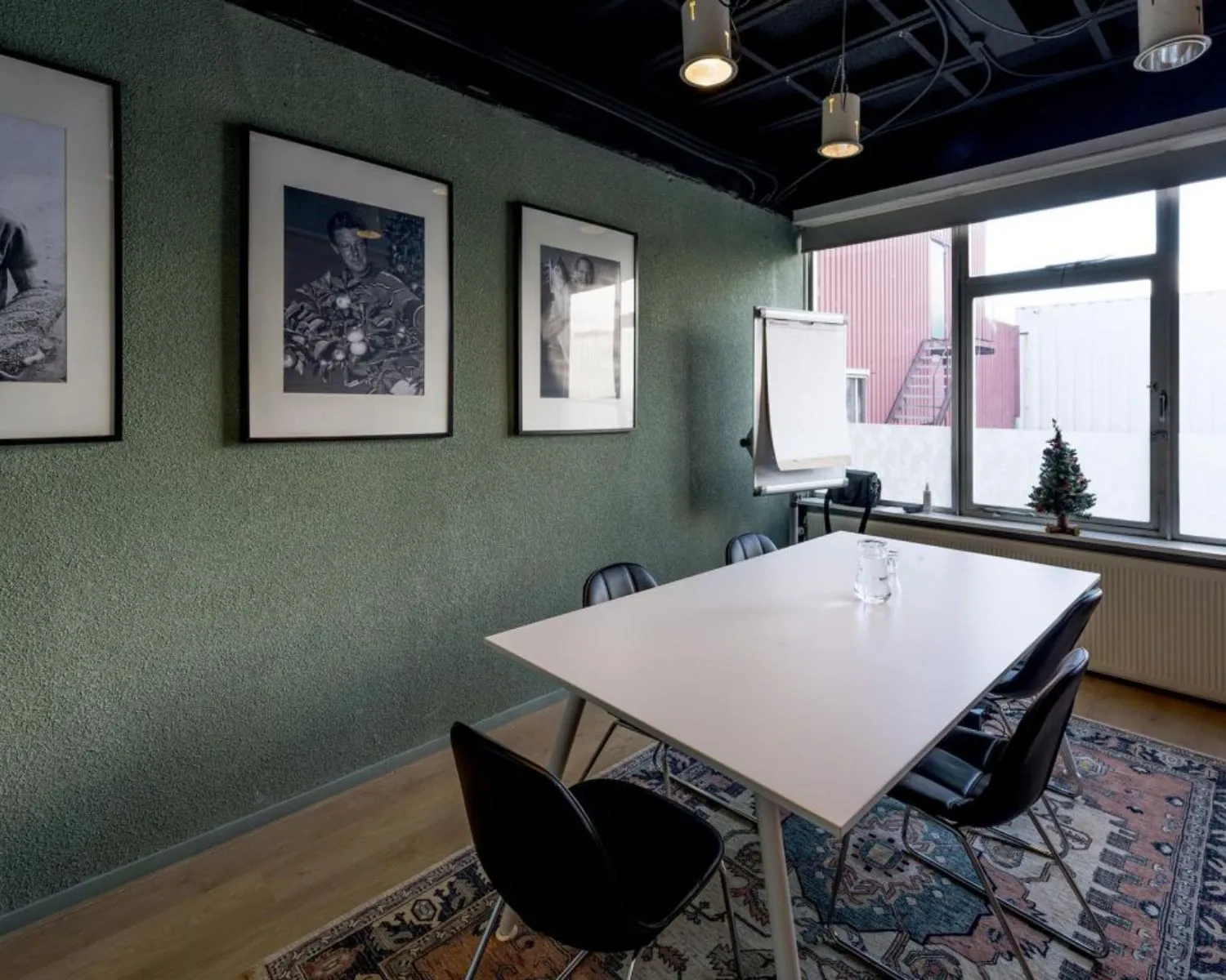 Kleine vergaderruimte met een witte tafel, zes zwarte stoelen, drie zwart-witfoto's aan de muur en een flip-over bij het raam.