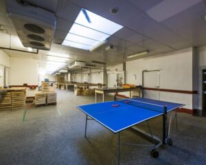 Een tafeltennistafel met rode batjes staat in een ruime, industriële werkruimte met houten kratten en tl-verlichting.