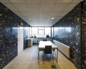 Vergaderruimte met donkere marmeren muren, een witte tafel met stoelen en een keukenblok bij het raam.