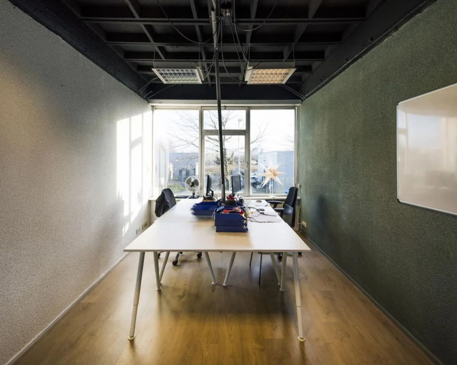 Kleine vergaderruimte met een lange tafel, vier stoelen, een whiteboard en een groot raam met zicht op buiten.