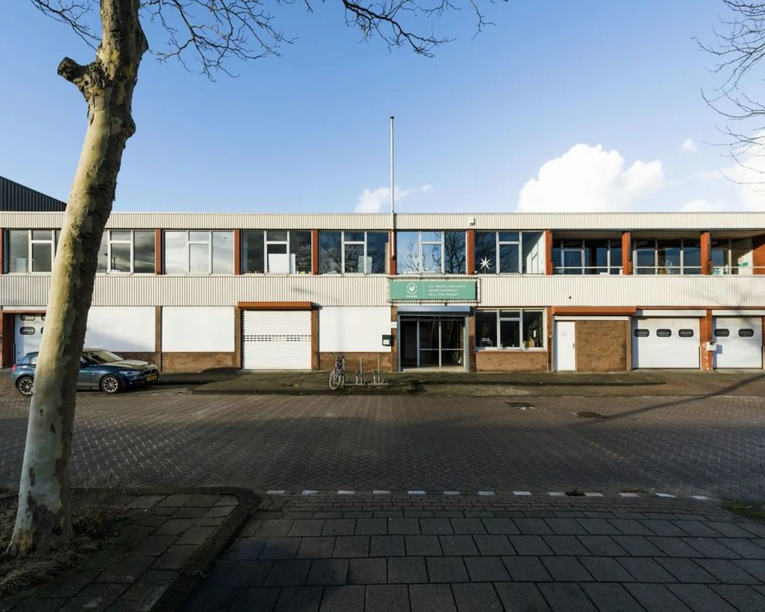 Vooraanzicht van een laag bedrijfsgebouw met witte gevel, grote ramen en een bord boven de entree.