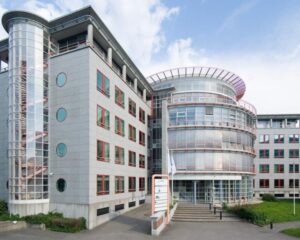 Modern kantoorgebouw met ronde glazen gevel en rode raamkozijnen onder een deels bewolkte lucht.