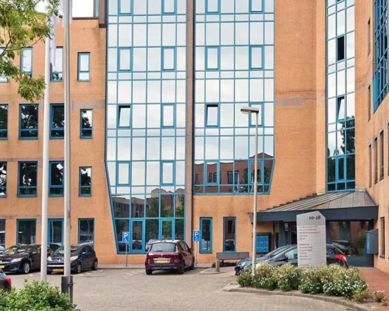 Vooraanzicht van een modern kantoorgebouw met spiegelende ramen en geparkeerde auto's op de parkeerplaats.