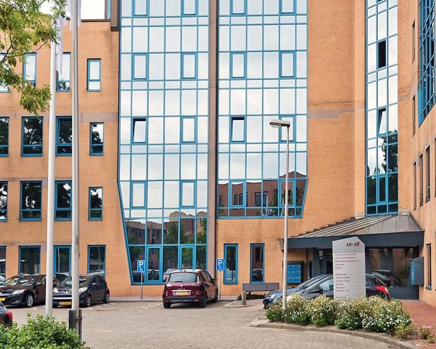 Vooraanzicht van een modern kantoorgebouw met spiegelende ramen en geparkeerde auto's op de parkeerplaats.