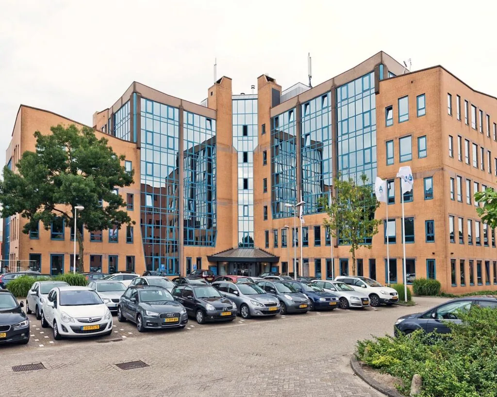 Kantoorgebouw met glazen ramen en een parkeerplaats vol auto's op de voorgrond.