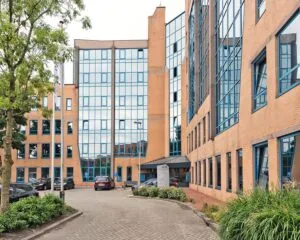 Ingang van een modern kantoorgebouw met oranje bakstenen en blauwomrande ramen, omringd door geparkeerde auto's en groenvoorziening.