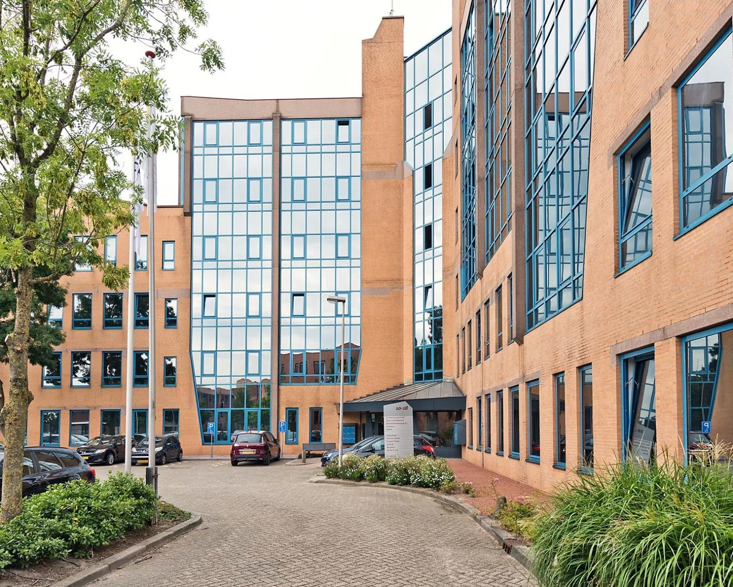 Ingang van een modern kantoorgebouw met oranje bakstenen en blauwomrande ramen, omringd door geparkeerde auto's en groenvoorziening.