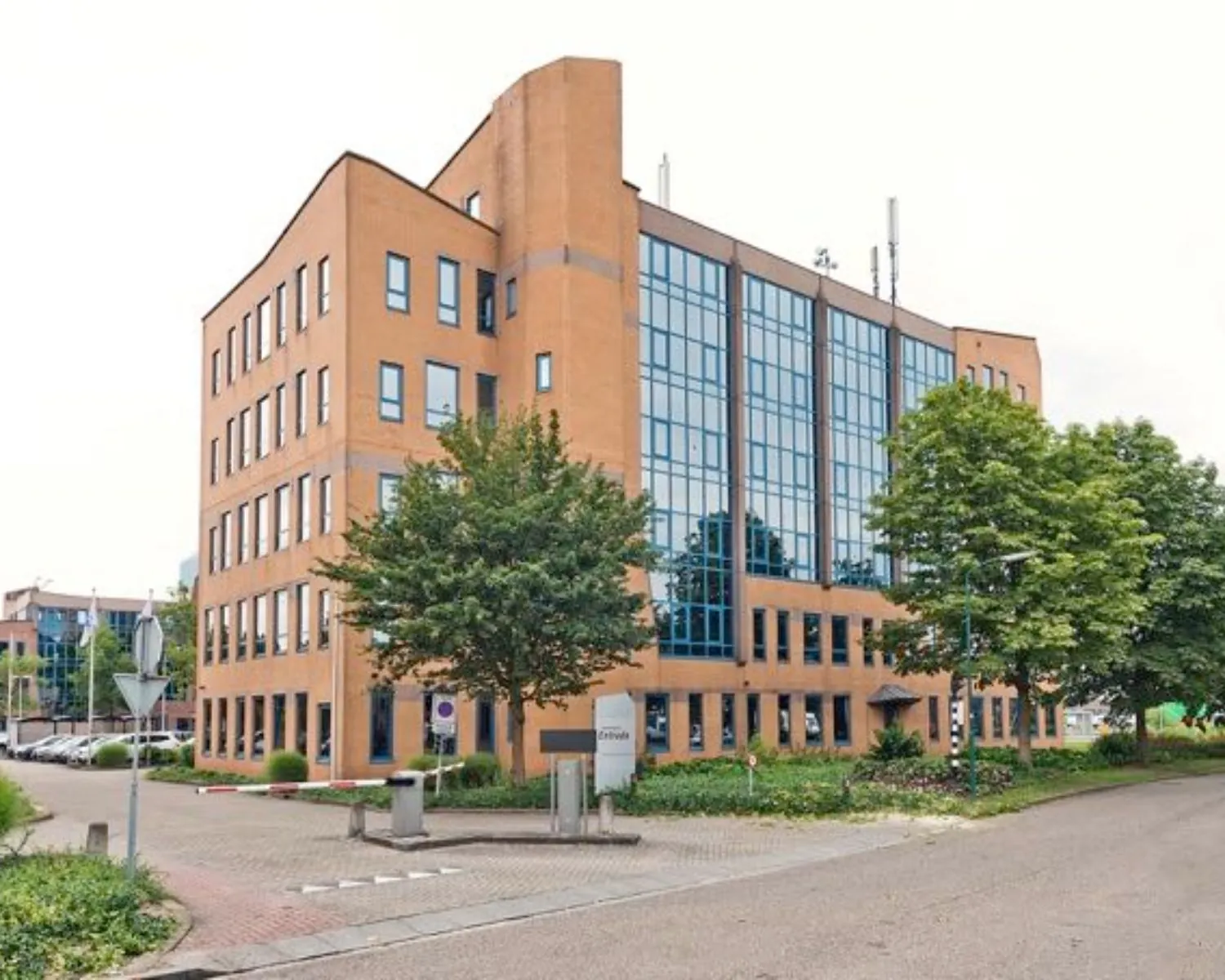 Bakstenen kantoorgebouw met grote blauwe ramen, omgeven door bomen en parkeerruimte.