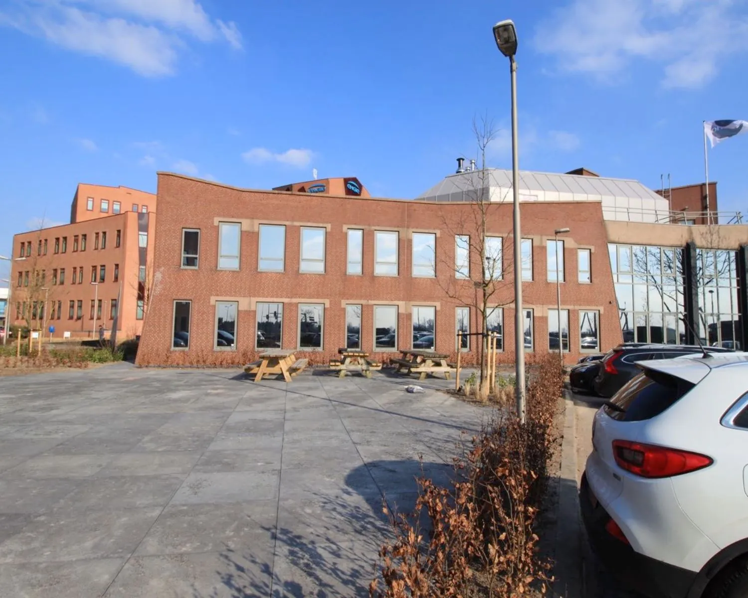 Modern kantoorgebouw met rode bakstenen gevel, parkeerplaatsen en picknicktafels op een ruim betegeld plein.