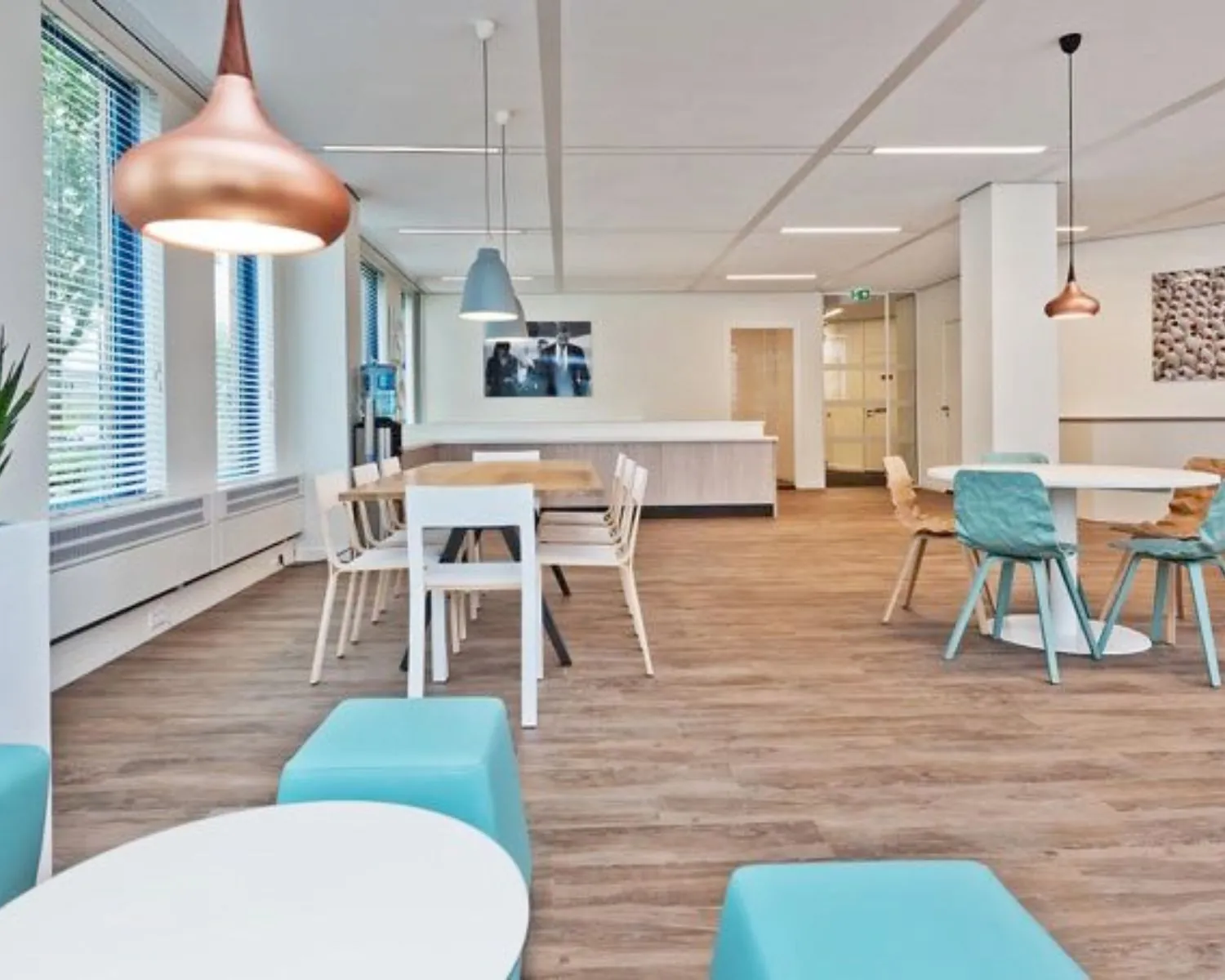 Moderne, licht ingerichte kantoorkantine met houten vloer, kleurrijke stoelen en hanglampen.