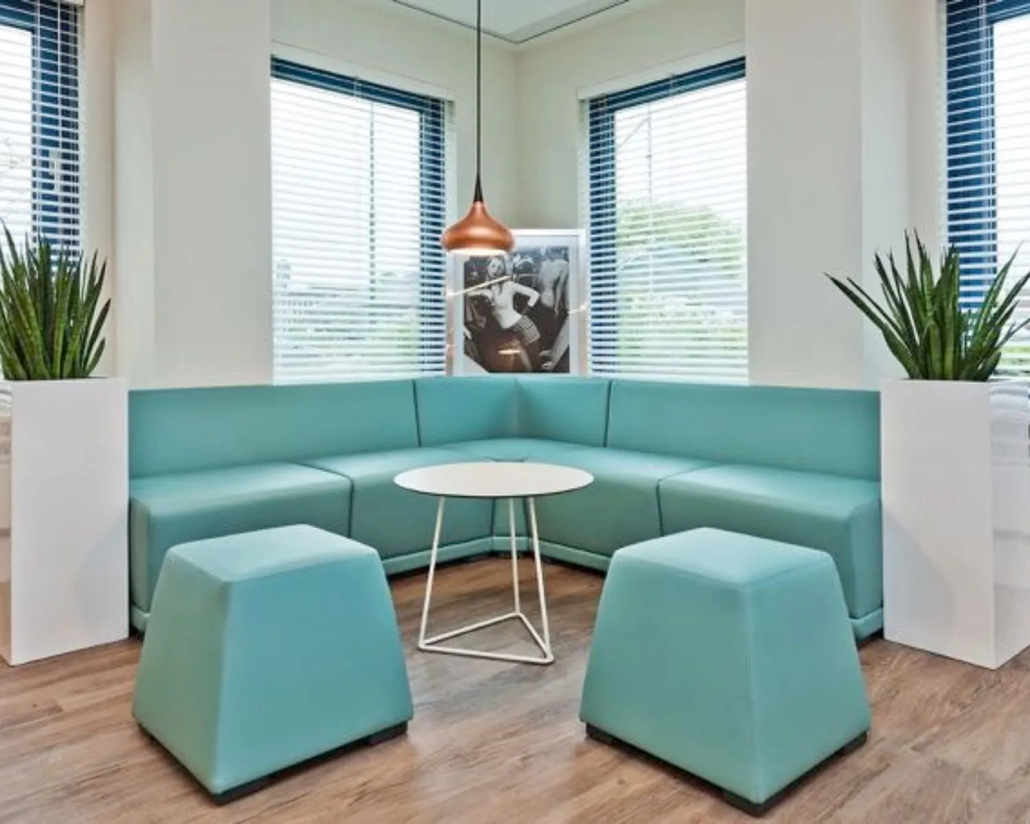 Wachtkamer met lichtblauwe hoekbank, moderne meubels, kamerplanten en een kunstfoto aan de muur.