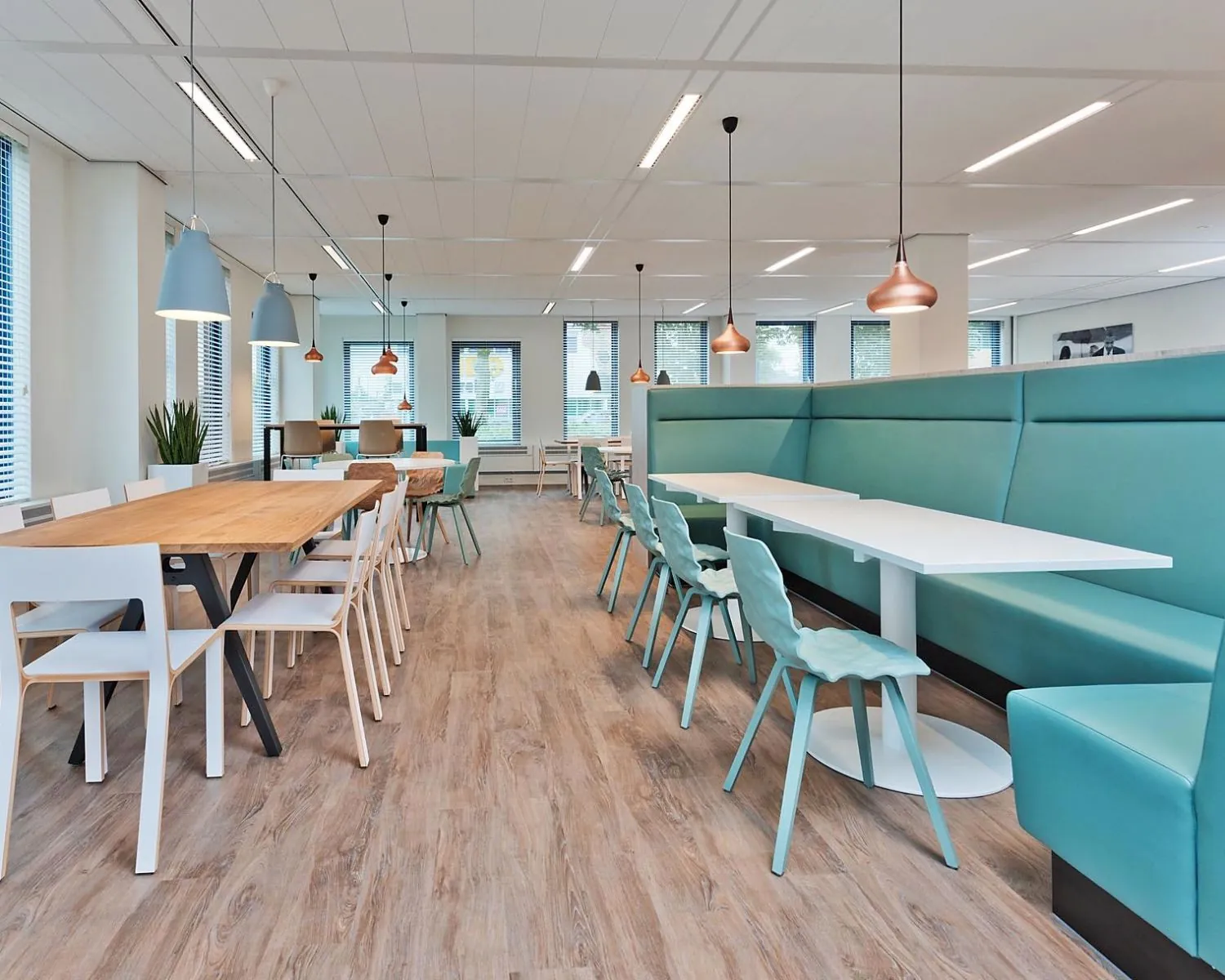 Modern ingerichte kantine met houten vloer, kleurrijke stoelen en hanglampen boven lange tafels.