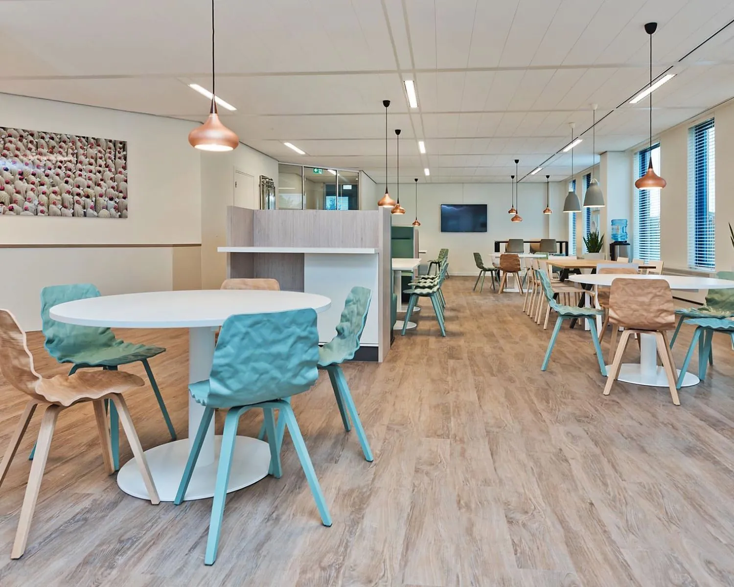 Moderne, open bedrijfsruimte met houten vloer, kleurrijke stoelen en ronde tafels, verlicht door hanglampen.