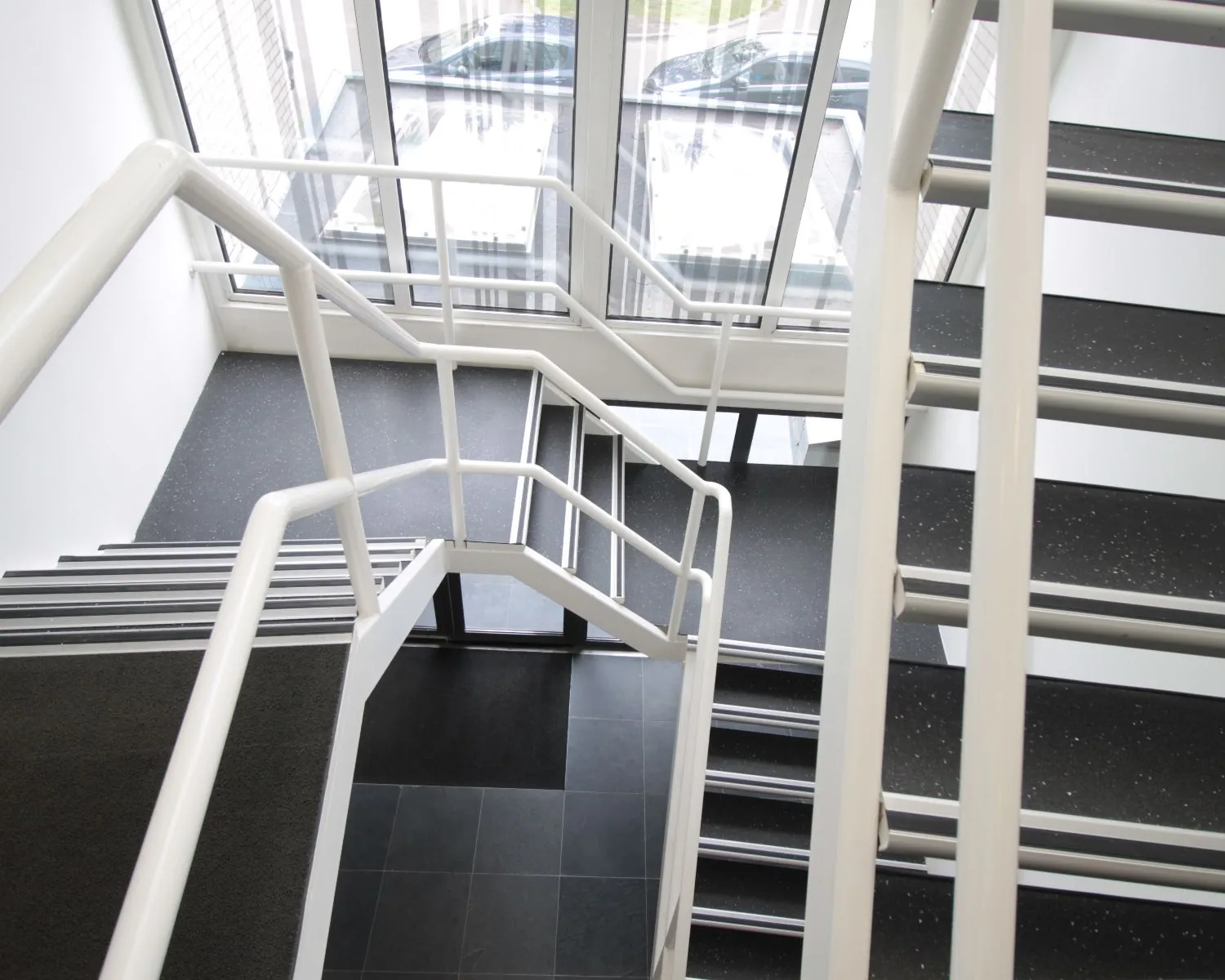 Trappenhuis met witte leuningen en zwarte traptreden in een modern gebouw met grote ramen.
