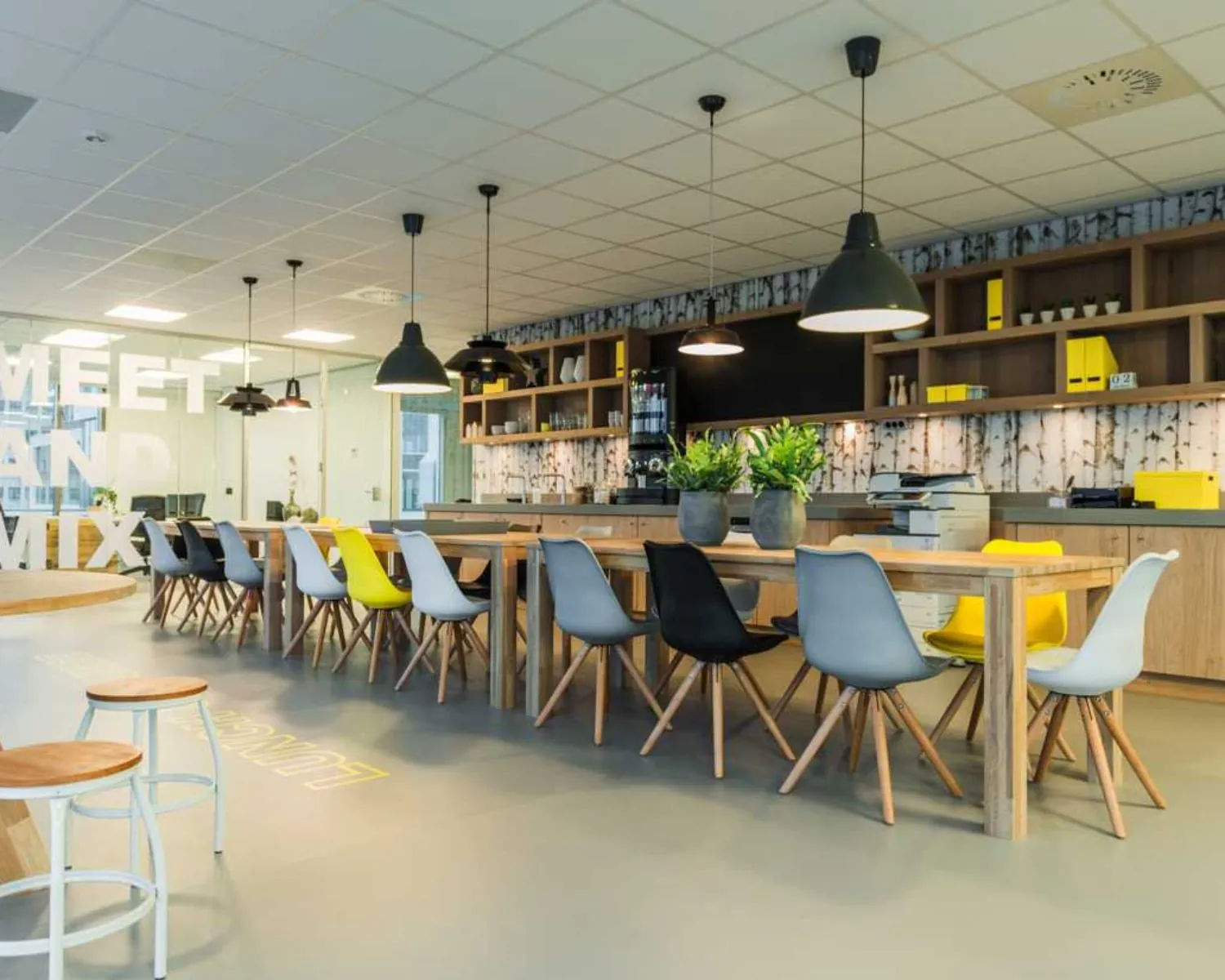 Moderne kantoorkantine met lange houten tafels, kleurrijke stoelen en hanglampen in een open en lichte ruimte.