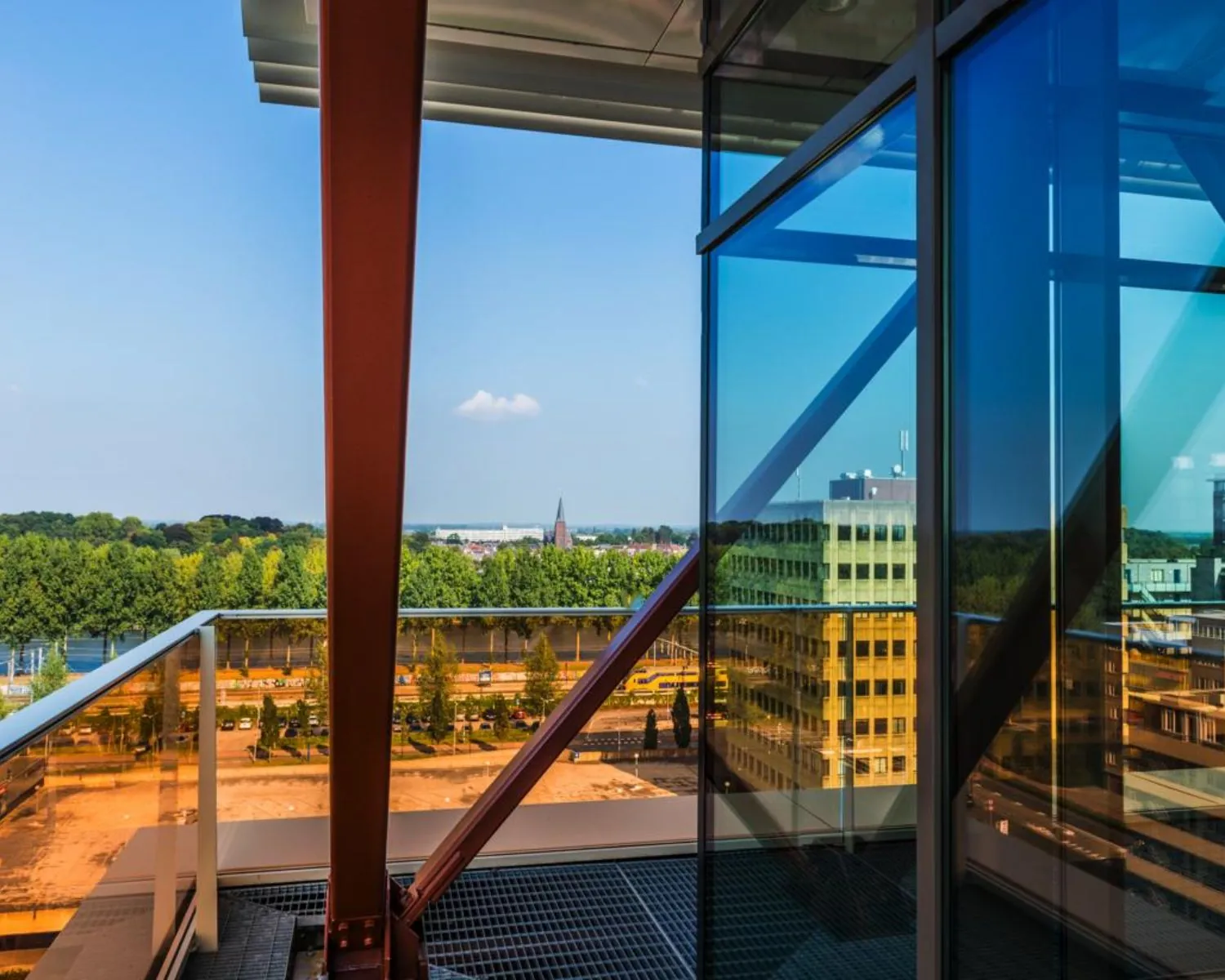 Uitzicht vanaf een modern glazen balkon met stalen constructie op een stad met groen, gebouwen en kerktoren in de verte.