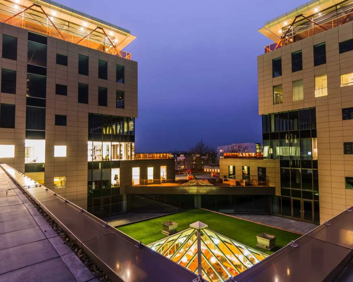 Twee moderne kantoorgebouwen met verlichte ramen en een glazen dakconstructie in het midden, gefotografeerd tijdens schemering.