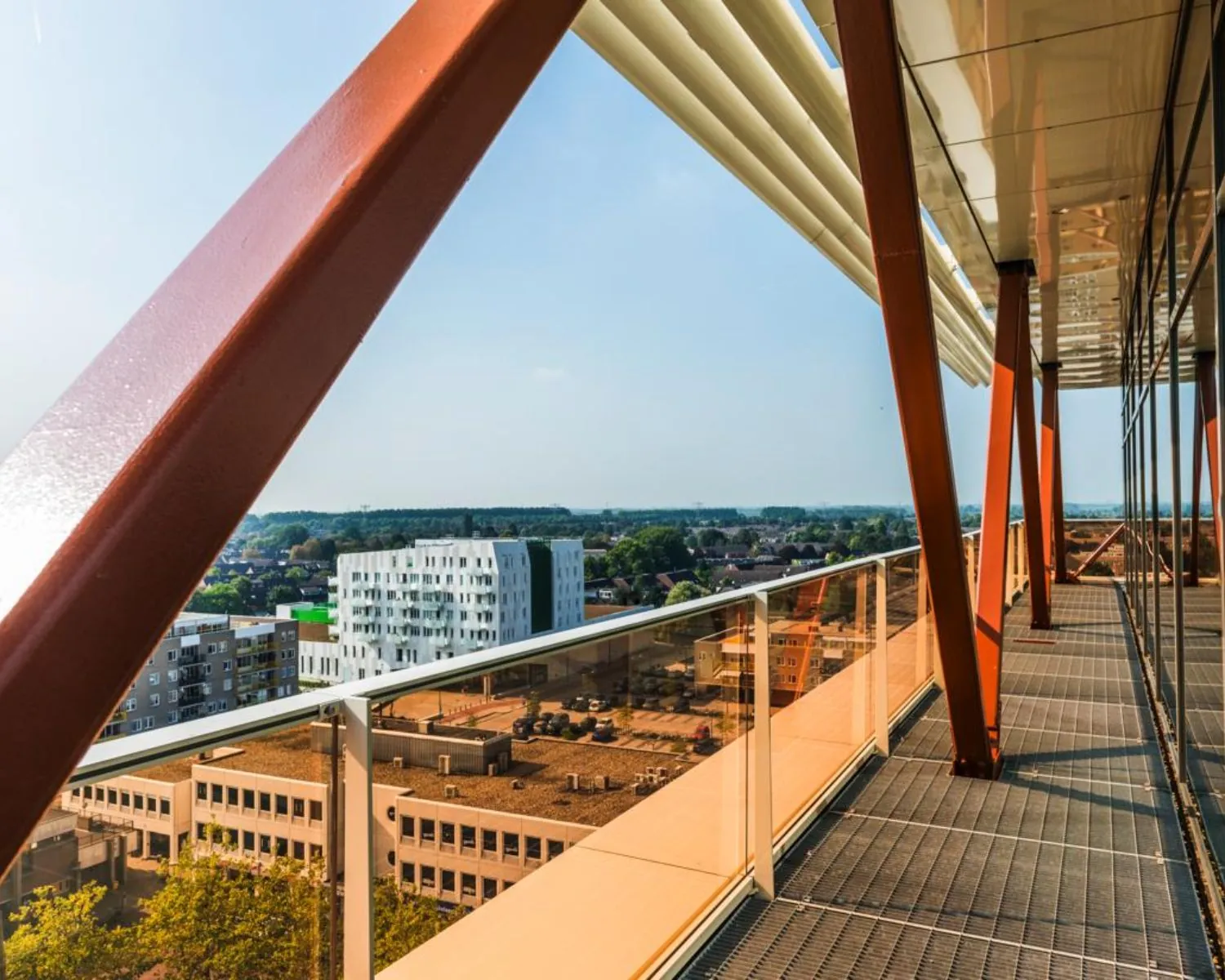 Uitzicht vanaf een hooggelegen balkon met oranje stalen balken en glazen reling over een stad met moderne gebouwen.