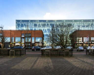 Straatbeeld met geparkeerde auto's, winkels (waaronder Zeeman) en een modern glazen kantoorgebouw achter een rij bomen.