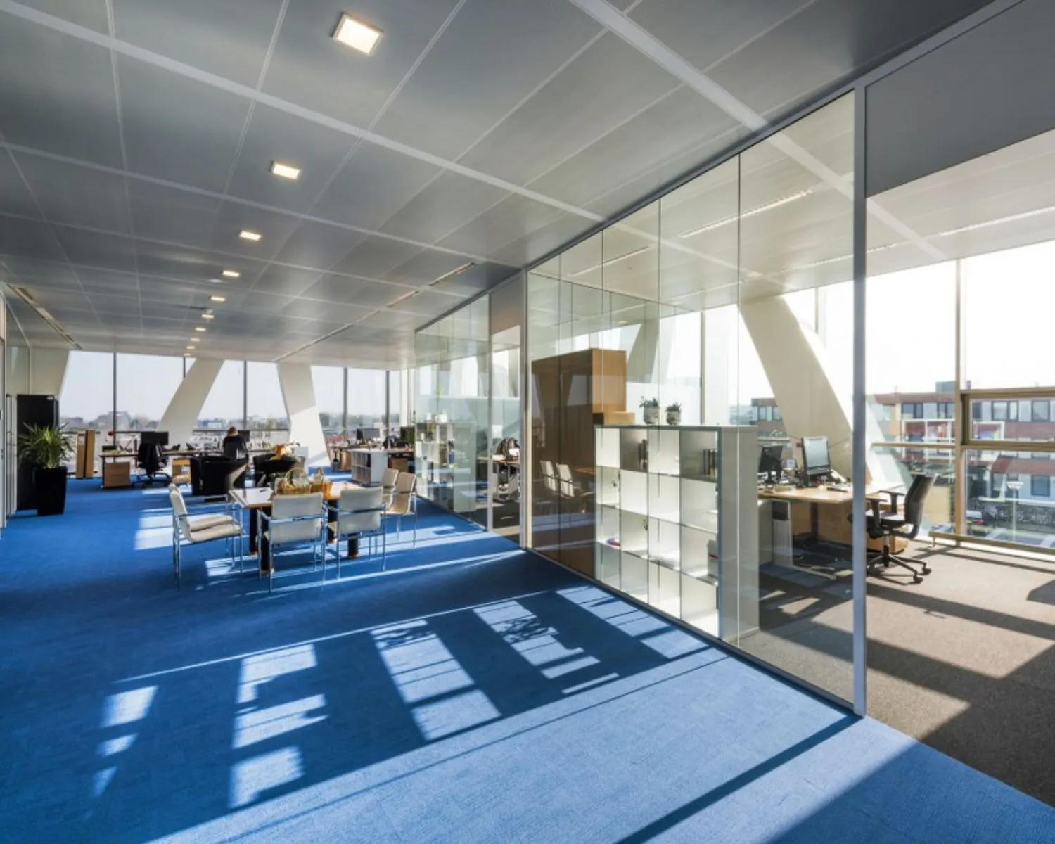 Modern open kantoorinterieur met glazen vergaderruimtes en blauwe vloerbedekking, verlicht door natuurlijk daglicht.