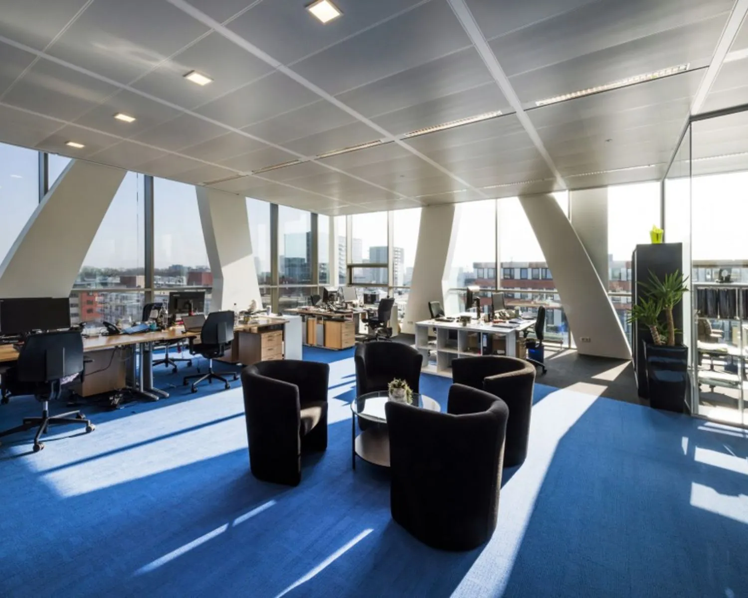 Modern kantoorinterieur met open werkplekken, grote ramen en een zitgedeelte met zwarte stoelen op blauwe vloerbedekking.