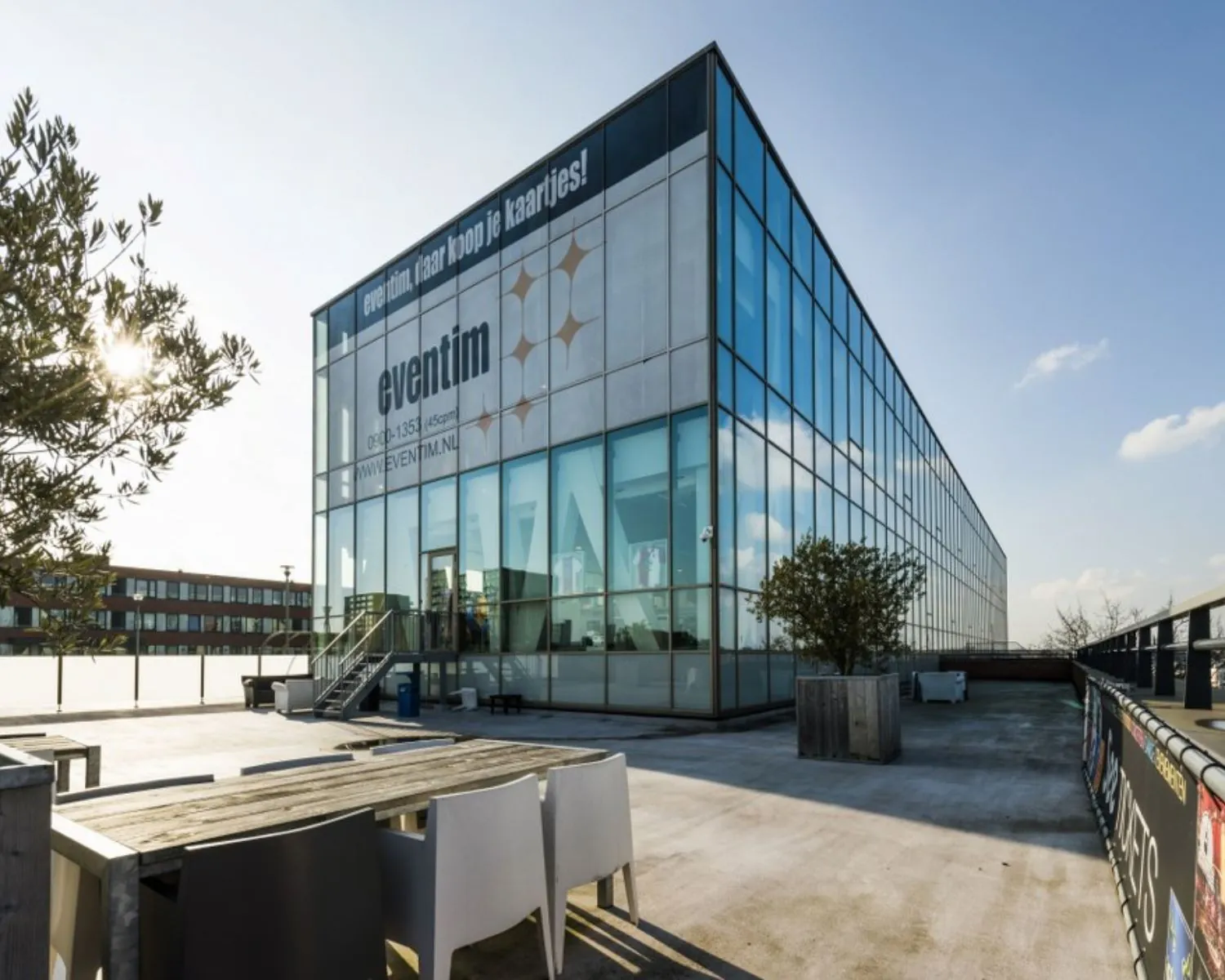 Glazen kantoorgebouw van Eventim met dakterras, picknicktafels en plantenbakken in de zon.
