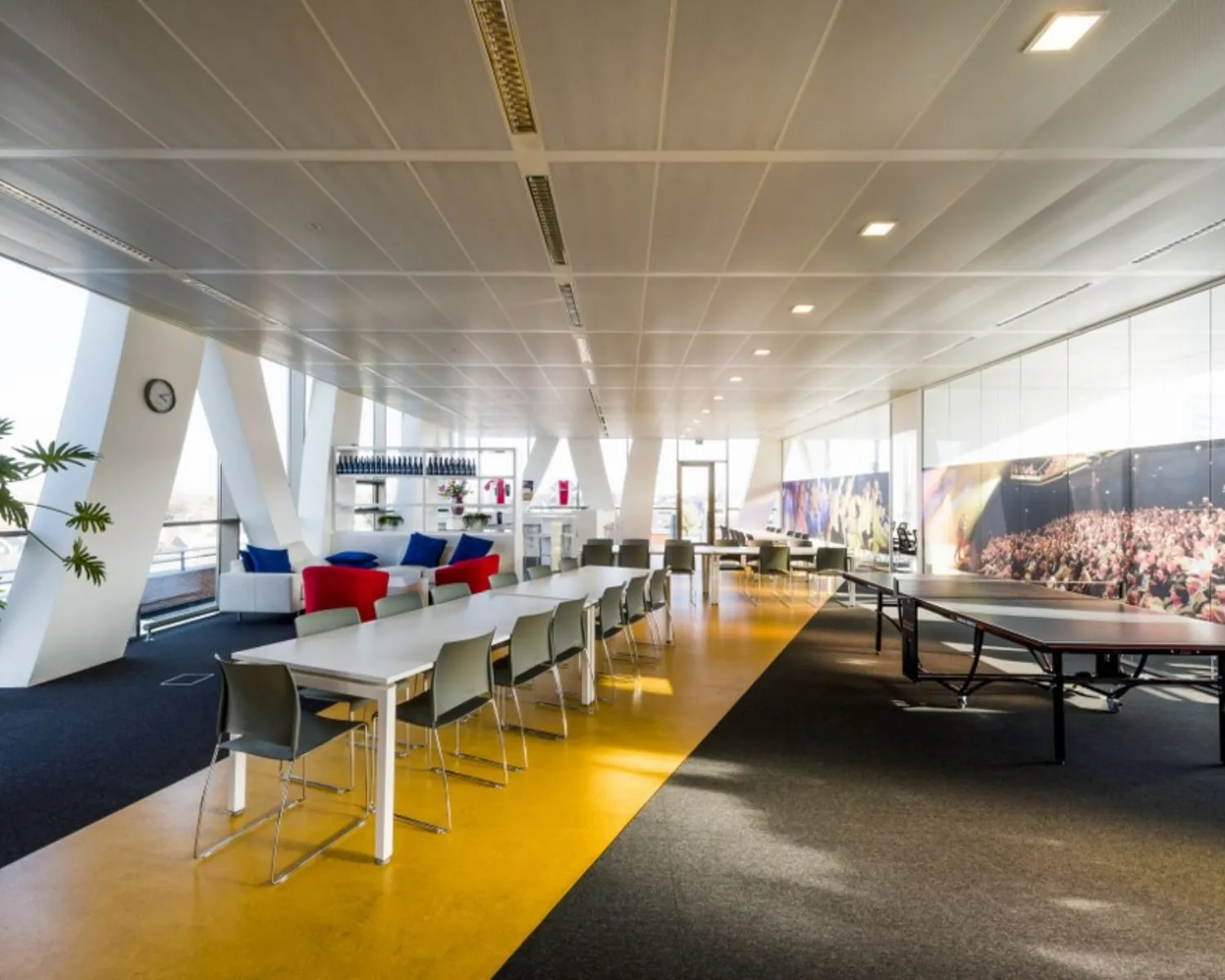Moderne en ruime kantoorlounge met zithoek, lange eettafels, een pingpongtafel en grote muurfoto van een concertpubliek.