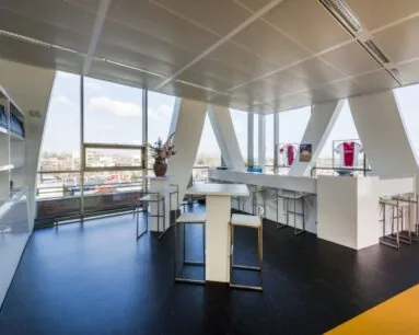 Moderne, lichte kantoorruimte met hoge ramen, statafels en vitrines met voetbalshirts.